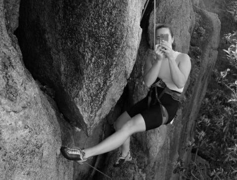 A woman rock climbing on Ko Tao, Thailand while capturing a photo with her smartphone.